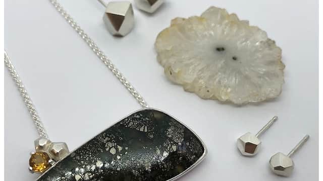 A silver necklace with a dark stone pendant, a yellow gem, and geometric silver earrings on a light background.