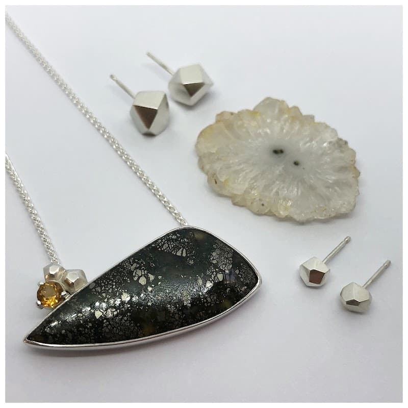 A silver necklace with a dark stone pendant, a yellow gem, and geometric silver earrings on a light background.