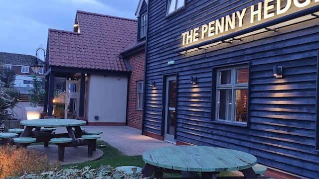 Exterior of The Penny Hedge bar with wooden benches, dark blue cladding, and a cloudy evening sky.
