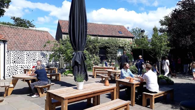 Outdoor seating area with wooden tables, a large umbrella, and greenery, bustling with diners on a sunny day.