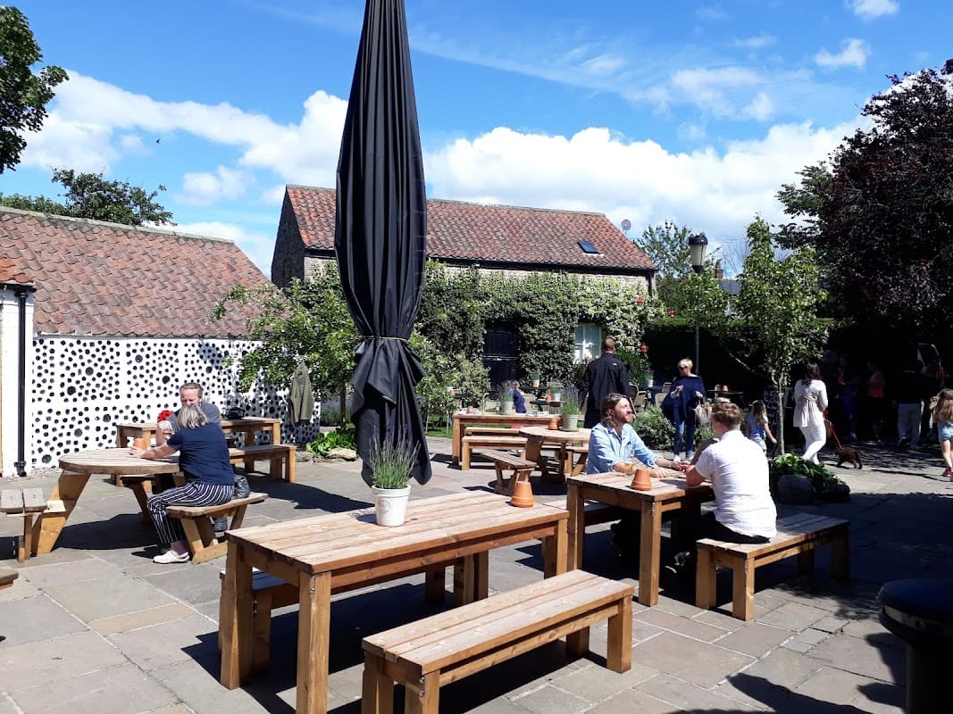 Outdoor seating area with wooden tables, a large umbrella, and greenery, bustling with diners on a sunny day.