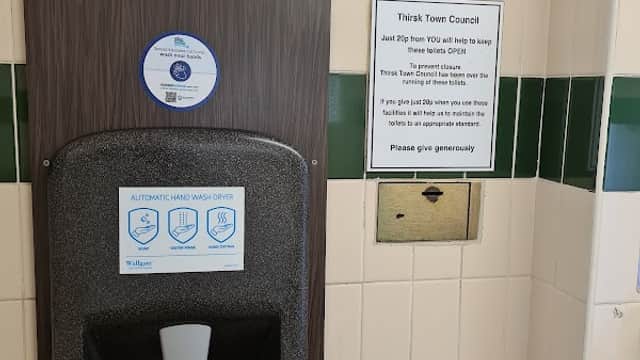 Automatic hand dryer mounted on a wall, with a sign about maintaining public toilets in Thirsk, Yorkshire.