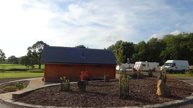 Wooden building with a circular garden, surrounded by green fields and parked caravans under a blue sky.