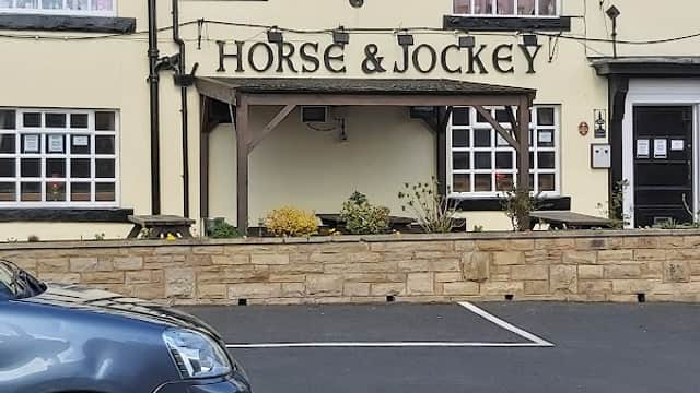 The facade of the Horse & Jockey pub in Birstall, featuring a sign and flower beds, with a parked car in front.
