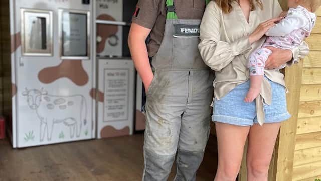 A man in overalls and a woman holding a baby stand outside a wooden shack with a milk vending machine.