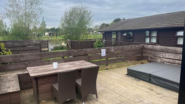 Wooden deck with a table and chairs, overlooking a tranquil pond and lodge, surrounded by greenery under a cloudy sky.
