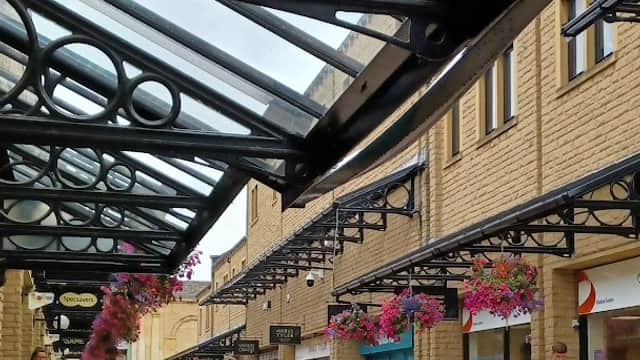 Covered walkway with shops, hanging flower baskets, and people walking in Woolshops Shopping Centre Car Park, Exley.