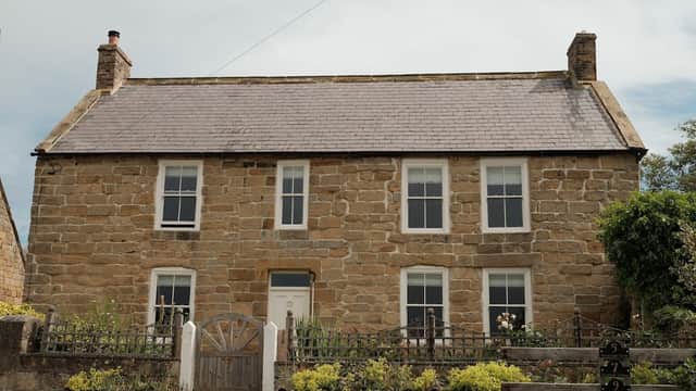 Stone cottage with a slate roof, white windows, and a wooden gate, surrounded by gardens in a rural setting.