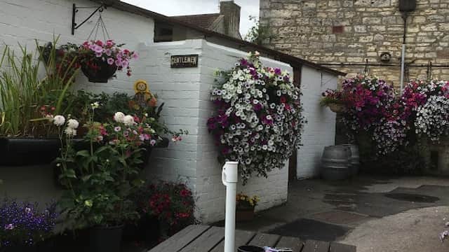 A rustic outdoor bar area with wooden tables, vibrant flower baskets, and a stone building in the background.