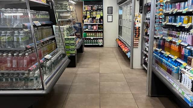A well-lit corner shop aisle with shelves stocked with drinks, snacks, and convenience items.