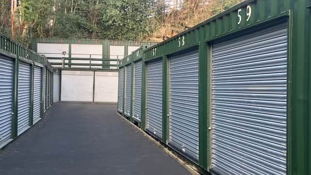 Green storage units with metal roll-up doors line a paved pathway, surrounded by trees in a quiet area.
