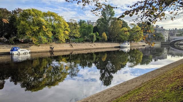 Pay & Display car park by a river, with trees and boats reflecting on the water under a blue sky.