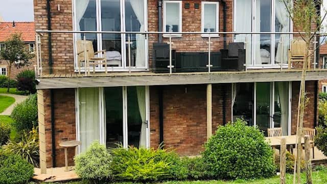 Two-story brick house with large windows, balcony seating, and a golden retriever lounging on green grass.