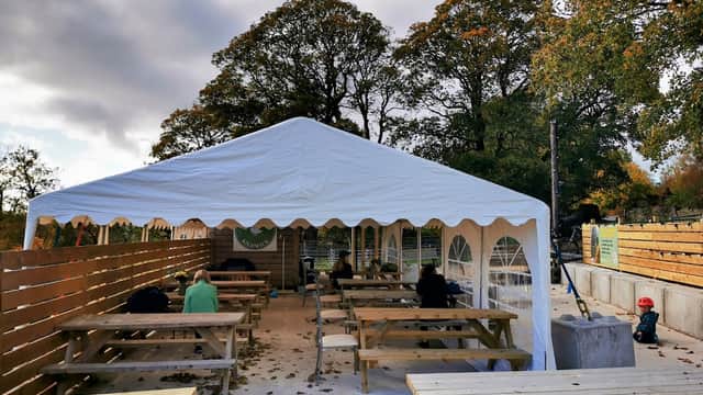 White tent with wooden benches, surrounded by trees, patrons seated, autumn leaves scattered on the ground.