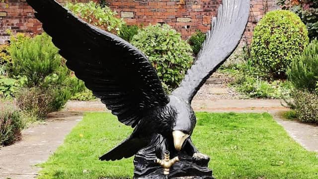 A large black eagle sculpture with outstretched wings perched on a rock, surrounded by greenery and a brick wall.