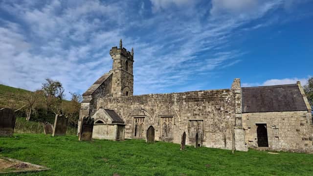 Medieval stone church with a tower, surrounded by grassy grounds and gravestones under a blue sky with clouds.