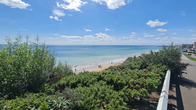 Scenic view of a beach with turquoise water, sandy shore, and people walking, framed by greenery and a clear blue sky.