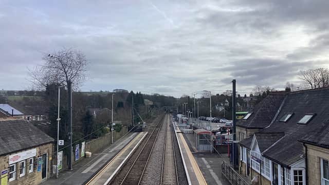 Train tracks stretch through Menston station, with buildings and trees lining the sides under a cloudy sky.