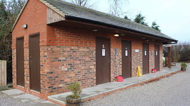 Red brick building with four doors, gravel path, and potted plants outside, set in a green landscape.