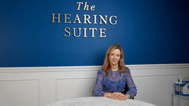 A woman in a blue blouse sits at a marble table in front of a blue wall with "The Hearing Suite" sign.