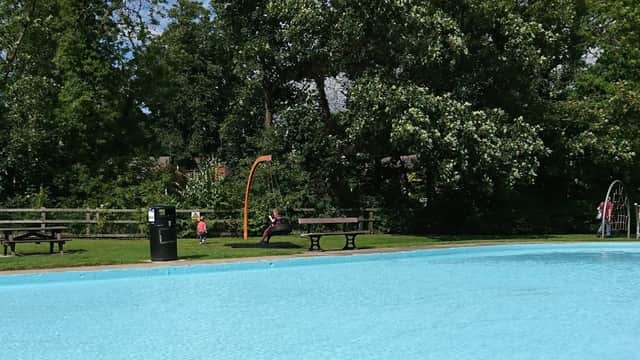 A clear paddling pool surrounded by green trees, benches, and a play area with a slide in Bridge Hewick, Yorkshire.