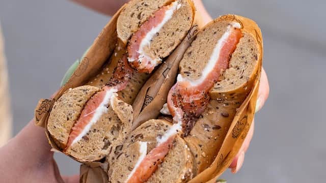 A person holding a halved bagel filled with cream cheese and smoked salmon, wrapped in paper.
