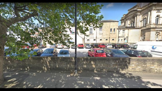 Parking lot in Huddersfield with various cars, trees, and historic buildings in the background.