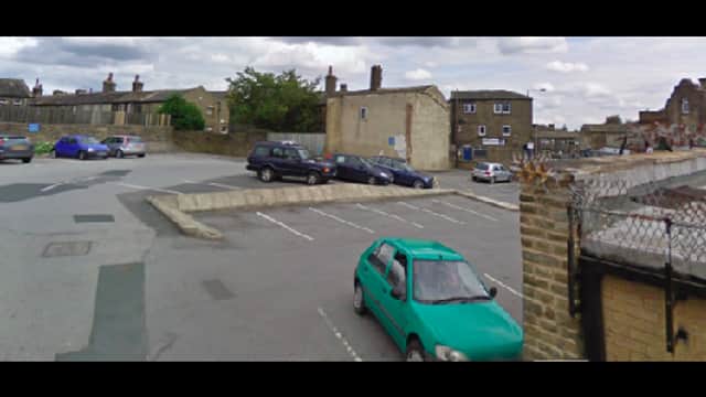 Car park with multiple vehicles, including a green car, surrounded by buildings in Shelf, Yorkshire.