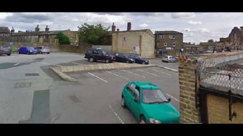 Car park with multiple vehicles, including a green car, surrounded by buildings in Shelf, Yorkshire.