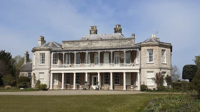 Historic building with a grand facade, large windows, and a balcony, set in landscaped grounds under a blue sky.