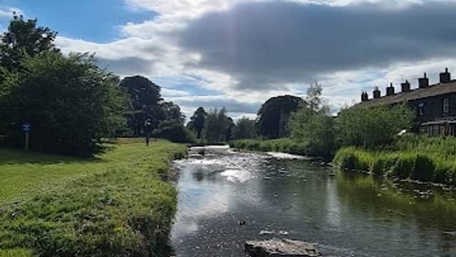 Sunlight reflects on a tranquil river, surrounded by greenery and stone, with buildings visible in the background.