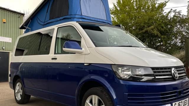 A blue and silver camper van with a pop-up roof parked outside a dealership, surrounded by greenery and cloudy skies.