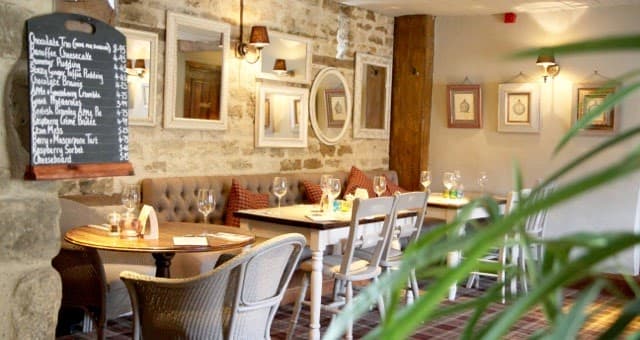 Cozy bar interior with stone walls, framed artwork, wooden tables, and a chalkboard menu. Greenery in the foreground.