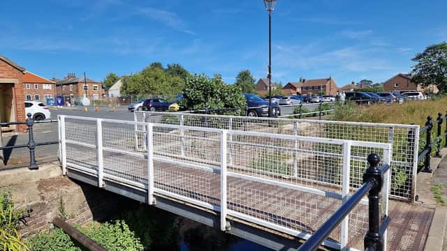 Bridge over a stream leading to Eastgate Free Car Park, with parked cars and grassy areas under a clear blue sky.