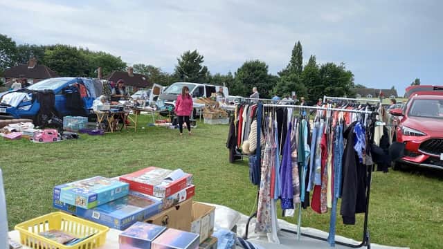 Vendors and shoppers browse various items at a busy car boot sale, with clothes hanging on a rack and parked cars nearby.
