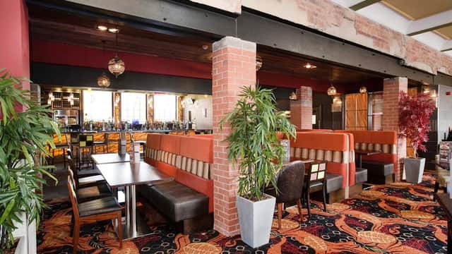 Interior of The Queens Hotel with vibrant carpet, cozy booths, and a bar in the background. Green plants add decor.