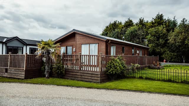 Wooden lodge with a deck, surrounded by greenery and a gravel path, set against a cloudy sky in Little Eden Country Park.