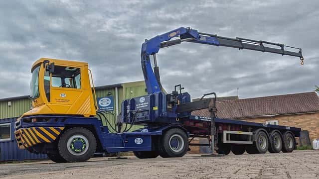 Yellow and blue truck with a crane attachment parked on a paved area, surrounded by industrial buildings and cloudy sky.