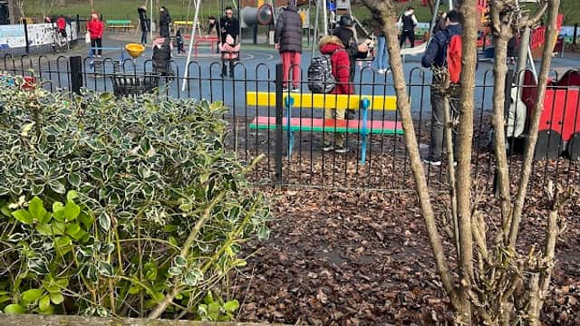 Lakeside park with a playground, children playing, and trees in a grassy area, surrounded by a black fence.