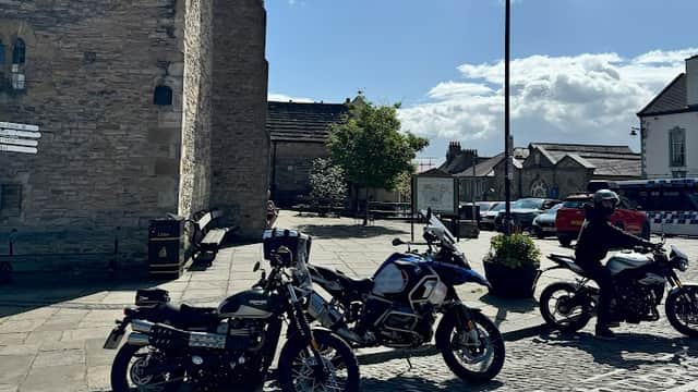 Motorcycles parked in a cobbled area with a pay and display sign, surrounded by buildings and a clear blue sky.
