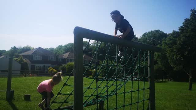 Children playing on a climbing frame in a sunny park, with green grass and houses in the background.