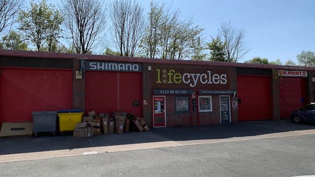 Red storefront with "Shimano" and "lifecycles" signage, surrounded by trees and parked cars. Boxes are stacked outside.