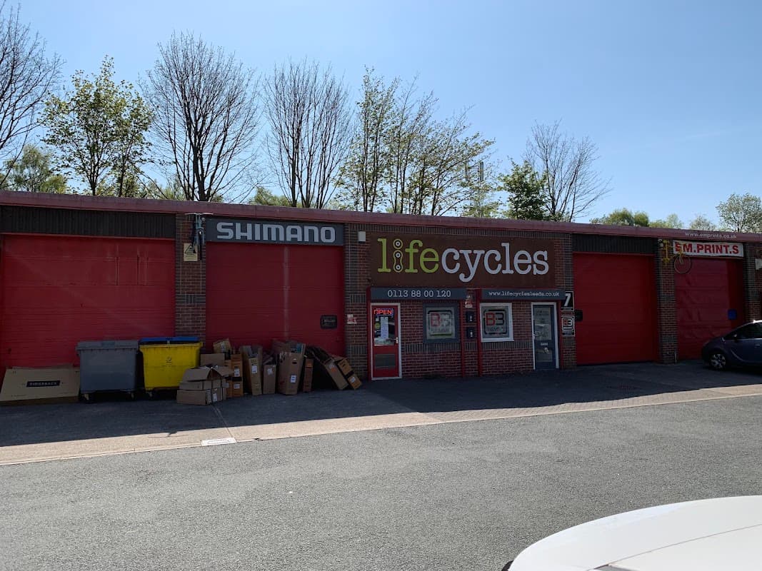 Red storefront with "Shimano" and "lifecycles" signage, surrounded by trees and parked cars. Boxes are stacked outside.