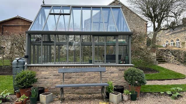 Glass greenhouse with stone base, surrounded by potted plants and a bench, set in a garden with stone buildings.