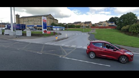 Red car driving on a road near a parking area and buildings in Wibsey, Yorkshire, with green grass and cloudy sky.