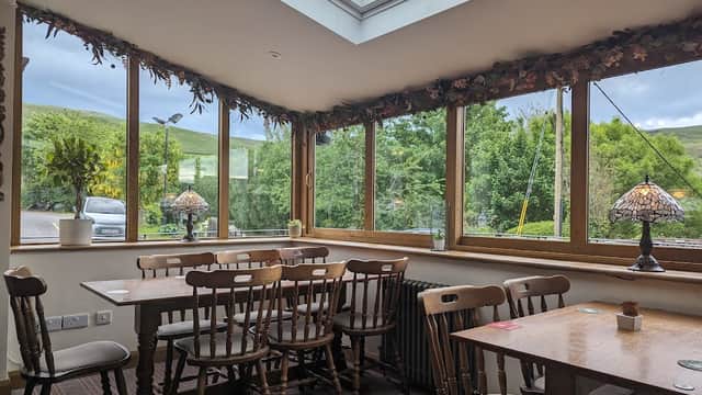 Cozy bar interior with wooden tables, chairs, and large windows overlooking lush greenery. Warm lighting and decor.