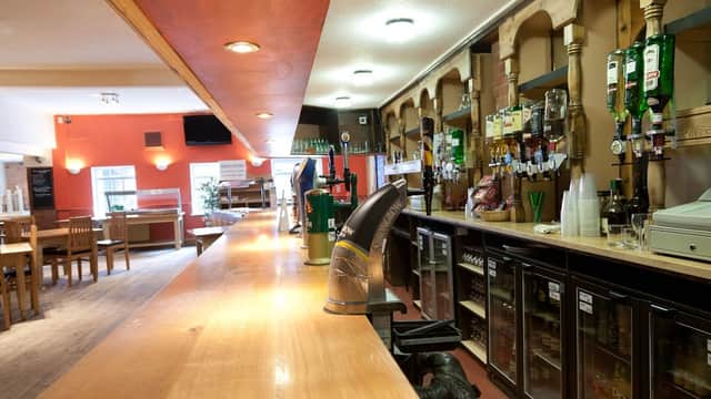 Wooden bar with drinks shelves, beer taps, and fridges; casual seating area in background with tables and chairs.
