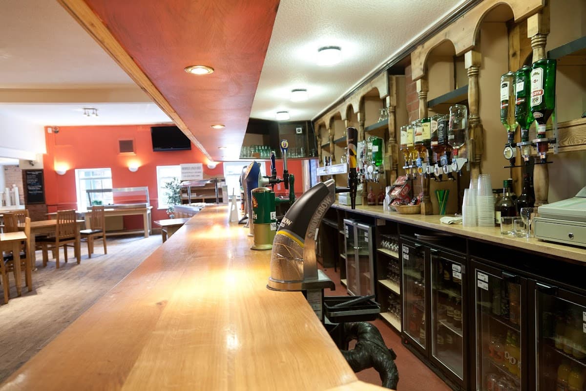 Wooden bar with drinks shelves, beer taps, and fridges; casual seating area in background with tables and chairs.