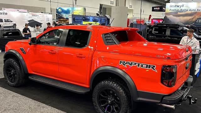 Bright orange Ford Raptor truck showcased at a car parts exhibition, featuring rugged tires and a sporty design.