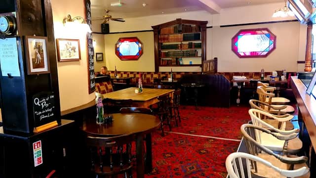 Cozy bar interior with wooden tables, chairs, patterned carpet, bookshelves, and stained glass windows.